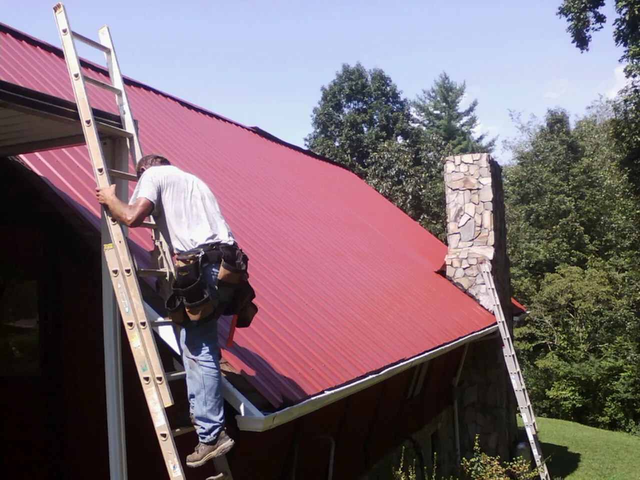Roofer on a ladder beside a red metal roof and stone chimney — Soffit & Fascia Repair by Roofing Brothers in Frenchburg